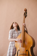 Beautiful young woman musician sitting on a vintage double bass on a beige background in a studio