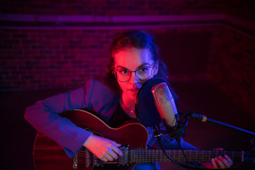 A young woman in glasses playing guitar and singing by the mic in neon lighting
