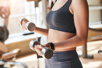 Close-up photo of young woman in sportswear and with smartwatch on her hand exercising with dumbbells while standing at gym
