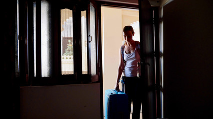 Silhouette of young woman in the hotel with a suitcase opens the door and passes to the dark room. View from the inside.