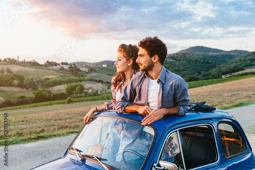 Loving couple on summer afternoon watching sunset from roof by old car around classical landscape of Tuscany, vineyards and farmland