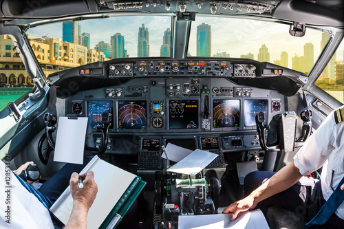 Poster Airplane cockpit flying on Dubai skyscrapers skyline, United Arab Emirates, with pilots arms and blank white papers for copy space