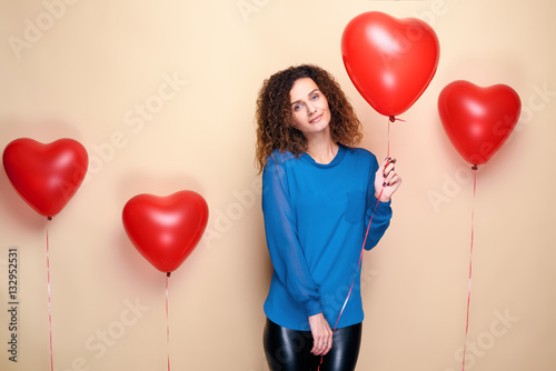 Poster Beautiful young girl with curly hair and blue sweater holding a few red heart air balloon and smiling