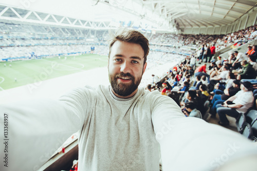 Poster Handsome bearded supporter watching football game and making selfie self-portrait with smartphone at Camp Nou, Barcelona, Spain