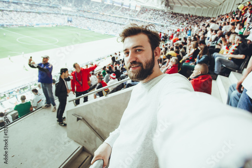 Poster Handsome bearded supporter watching football game and making selfie self-portrait with smartphone at Camp Nou, Barcelona, Spain