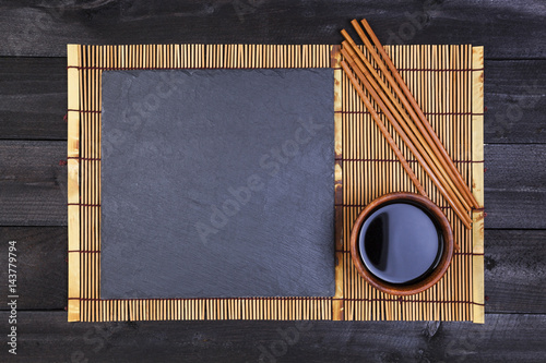 Background for sushi. Bamboo mat and soy sauce on black wooden table. Top view with copy space