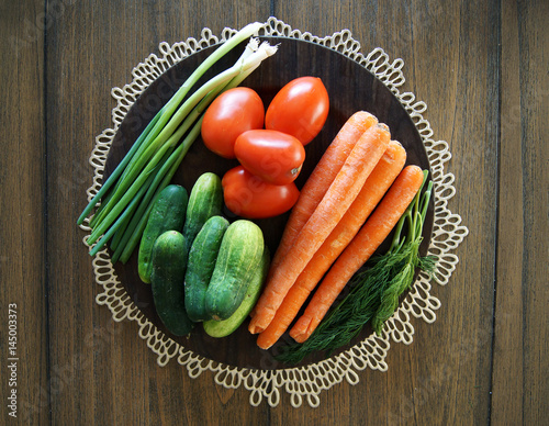 Colorful close up photo of vegetable plate with cucumbers, carrots, oval tomatoes, dill and long white bunching evergreen onion from garden on the dark wood finish table © Iryna