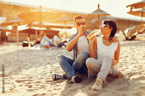 Happy young couple drinking beer and having fun at the beach during sunset