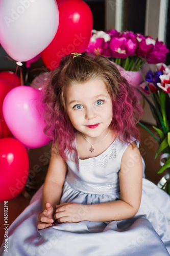 Poster Little beautiful sweet girl with pink curly hair in a silver dress in red tulips and with multi-colored balls smiling and posing
