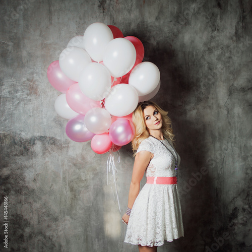 Poster Charming young blonde in a white dress with pink sash, holding a large bundle of balloons at the party