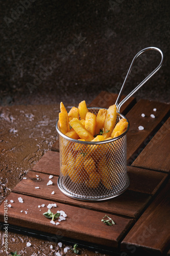 Poster Traditional french fries potatoes served in frying basket with salt, thyme on wooden board over brown texture background