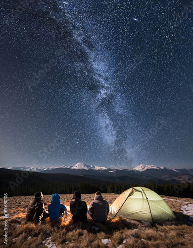 Rear view of four people sitting together beside camp and tent under beautiful night sky full of stars and milky way. On the background snow-covered mountains. Long exposure