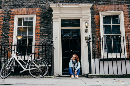 Obraz na płótnie .Young woman using her cellphone, seated infront of a door in an autumn day on the street of London. Lifestyle Photography