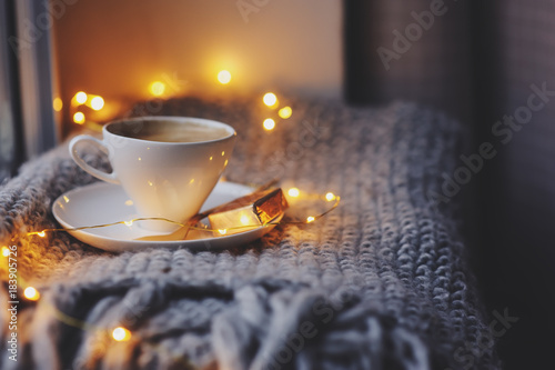 cozy winter or autumn morning at home. Hot coffee with gold metallic spoon, warm blanket, garland and candle lights, swedish hygge concept.