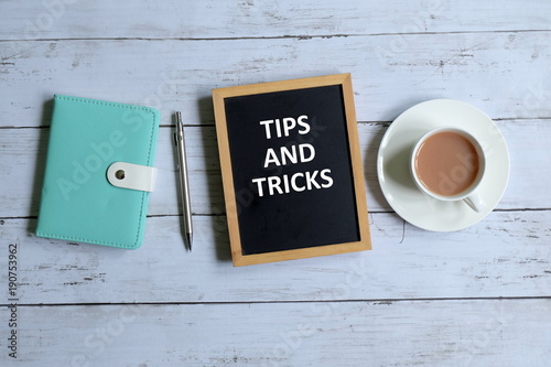 Top view of notebook,pen,a cup of coffee, and blackboard written with 'TIPS AND TRICKS' on white wooden background.