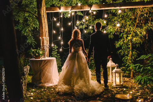 Night wedding ceremony with candles, lanterns and lamps on tree. Bride and groom holding hands on background of baulb lights, back view. Beautiful young couple standing under a tree at night