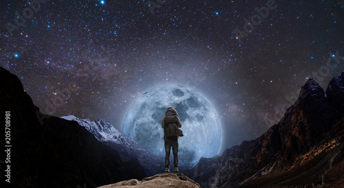 a man with backpack standing on mountain peak at night, and silhouette mountain with bright full moon and sky full of stars