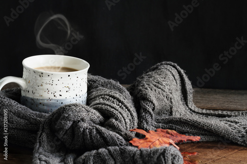 Hot steaming cup of coffee with cream wrapped in a cozy grey scarf with fallen leaves for autumn. Extreme shallow depth of field with selective focus on mug of coffee.