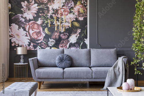 Blanket on grey couch in living room interior with flowers wallpaper and lamp on table. Real photo © Photographee.eu