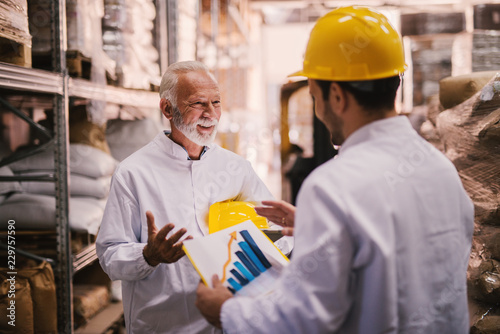 Picture of two male warehouse employees talking about trade success of their storage. Standing in warehouse in white clothes and yellow helmets . One of them is holding paper with histogram on it.