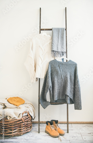 Fall or winter warm knitwear on hanger, white wall background. Fashionable female winter clothing. Woolen sweaters, grey scarf, boots and knitted hat