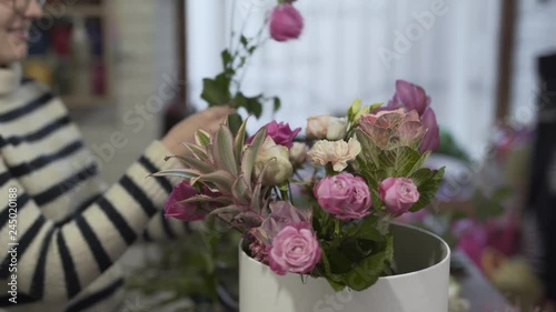 Female florist in a flower shop is making a bouquet in a round box from violet and purple kind of roses. Florist is wearing a round glasses and adding a new flowers to end a complete bouquet. © blackboxguild