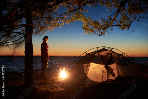 Night camping at sea coast under clear blue sky. Attractive tourist woman having a rest alone at campfire near tent on clear water and distant lights on horizon background. Tourism and recreation.