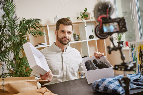 Fashion blog. Smiling male blogger opening box with sneakers while recording new video at home on a digital camera.