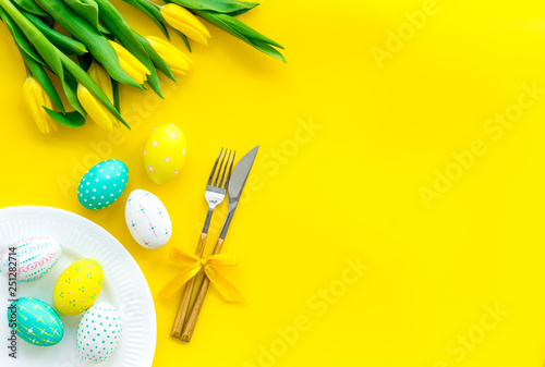Festive Easter table decorated with tulips. Tableware and painted eggs on yellow background top view copy space © 9dreamstudio