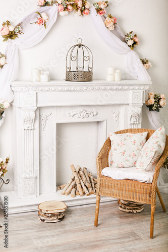 Wicker chair by a white decorative fireplace with firewood in a light interior © Semenova Jenny