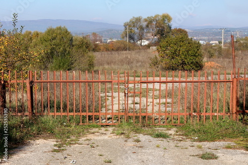 Long metal fence doors locked with chain and padlock preventing entrance to large abandoned industrial complex surrounded with high uncut grass and trees on clear blue sky background © hecos