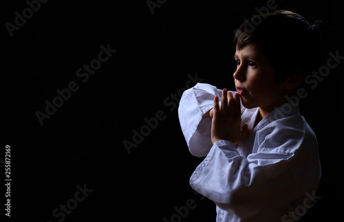 Little karate kid in white kimono isolated on dark background. Portrait of boy ready for martial arts fight. Child fighting at judo training. Best concept for martial fights and sport activity. © vmargineanu