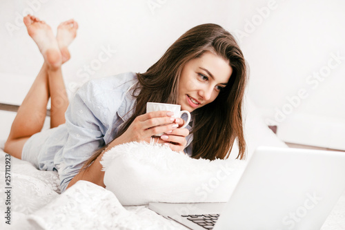 Young smiling woman working on laptop in her bedroom. Happy morning.