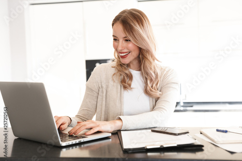 Beautiful blonde woman posing sitting indoors at home using laptop computer. © Drobot Dean