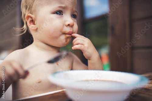 Small girl standing by a table on a patio in summer, eating soup. © Halfpoint
