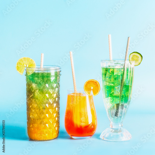 Various colorful summer refreshing lemonade long drinks with ice cubes and cocktails in glasses with paper drinking straws and citrus fruits at blue background © VICUSCHKA