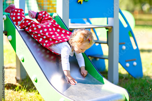 Cute toddler girl playing on slide on outdoor playground. Beautiful baby in red gum trousers having fun on sunny warm summer day. Child sliding down © Irina Schmidt
