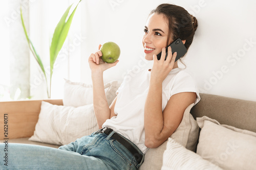 Beautiful happy young student girl sitting indoors eat apple talking mobile phone on sofa. © Drobot Dean