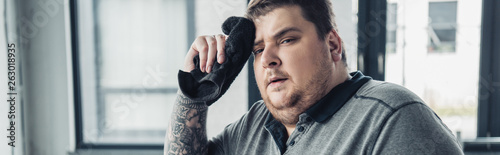 panoramic shot of tired overweight man looking at camera and wiping face with towel at sports center © LIGHTFIELD STUDIOS