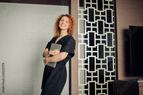 Positive caucasian female freelance blogger with frizzy red hair glad to participate in intresting meeting, posing at hotel with laptop. © alfa27
