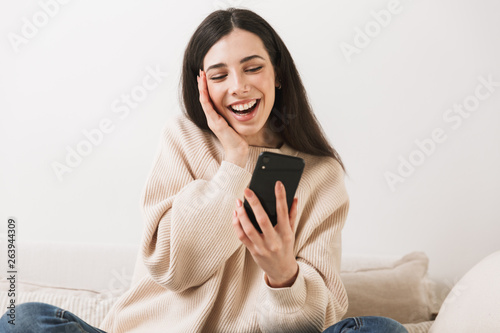 Photo of attractive caucasian woman holding cell phone while sitting on sofa at home © Drobot Dean